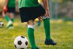 A boy wearing a green and black soccer uniform reaches down to hold his knee, a soccer ball next to him on a field.  