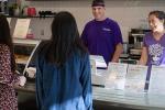 Two customers place their orders at a café. Two employees clad in purple T-shirts chat with them from behind the register. 