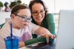 A mother and her son, who has Down syndrome, are sitting at a table working on a computer both wearing glasses