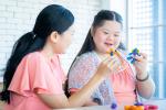 A mother with long black hair and daughter with long black hair, who has down syndrome, sit at a table playing with colorful plastic toys. 