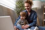 A mother and son sit on a couch, looking at a laptop, which is open beside them, as they engage with someone on a video call.  