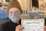 Woman with a tan knit hat and wrapped in a blanket and holding a certificate.
