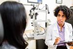 A female patient sits in a chair to undergo an eye exam with a Black female physician in a white coat and holding a clipboard.