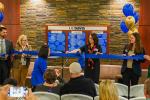 Woman in a blue blouse uses giant gold scissors to cut a blue ceremonial ribbon held by colleagues.