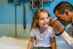 Doctor looks in girl’s ear while she smiles on patient table.  