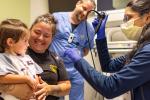 A preschool-aged girl sits on her mother’s lap in a patient room while a long-haired female doctor shows her the laryngosocopy scope with a lighted ti