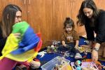 Two woman and a young girl play with colorful toys while seated on a colorful rug at a library playgroup
