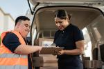 Two postal service employees, one a male with developmental disabilities, and one a female, stand together at the back of a white van filled with mail