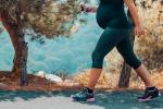 A pregnant woman walks outdoors, with pine trees in the background.