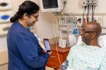 Woman in blue medical scrubs holds tablet while looking at patient lying in hospital bed.