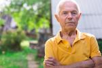An older man stands outdoors with his arms crossed, looking directly at the camera in a green, rural setting.
