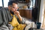Sick man on sofa coughing, wrapped in blanket, surrounded by tissues and toilet paper, feeling unwell.
