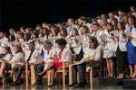 Group of people standing on stage wearing white coats and reading from booklet.