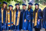 Eight graduates in commencement cap and gowns stand next to each other outside and smile at camera