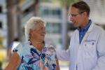 A person in a white coat speaking with someone in a floral shirt using a walker.