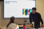 A woman, seated, talks to a man, standing, in front of a whiteboard containing a presentation in Spanish. 