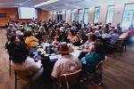 A conference room filled with people sitting around tables and directing their attention to guest speakers in the front of the room