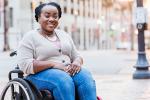 A mid adult Black woman in her 30s in a wheelchair outdoors, in the city, smiling at the camera. She has spina bifida.