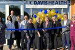 11 people stand outside the front doors of a clinic, with one man holding scissors to cut the blue ribbon in front of them