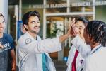 Two medical students in white coats high-five each other while other students look on, smiling, in front of library building 