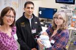 Female and male doctors in casual clothes stand next to a mother holding her infant baby in a hospital room.