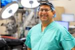 A surgeon wearing green scrubs, a cap and an augmented reality headset stands in an operating room.