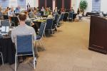 A woman at a podium speaks in a conference room where dozens of people are seated at round tables