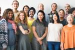 A diverse group of people stands in front of a white wall. 