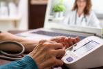 Close up of elderly hands using blood pressure machine at home while physician looks on in the background from a computer laptop screen