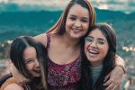 Three young women laugh as they pose for a photo, with arms around each other. They’re outside, on a hill, with city lights behind them. 