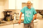 A man stands in his kitchen wearing a blue shirt that says "survivor"