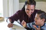 A woman sits at a table with a young boy who has Down syndrome. They are working with paper and crayons. 