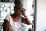 A mature Black woman is seated and holding her head with her left hand as if experiencing pain. 