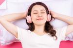 A young Asian woman with her eyes closed and wearing pink headphones is resting on a white pillow against a pink background.