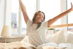 A smiling Black woman is sitting up in bed with her arms outstretched. 