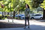 Woman with dark hair, wearing green sweatshirt and navy pants, walks down sidewalk with trees and park bench in background.