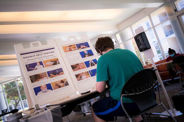 Photo of UC student sitting in front of a COVID-19 self testing station with instruction board. Credit: Erik Jepsen/UC San Diego