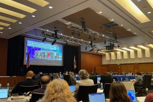 Dr. Carrie Byington at the speakers table in the UC Regents meeting room with regents sitting on the dias