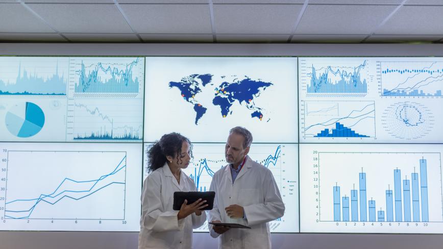 two researchers in white coats standing in front screens presenting data
