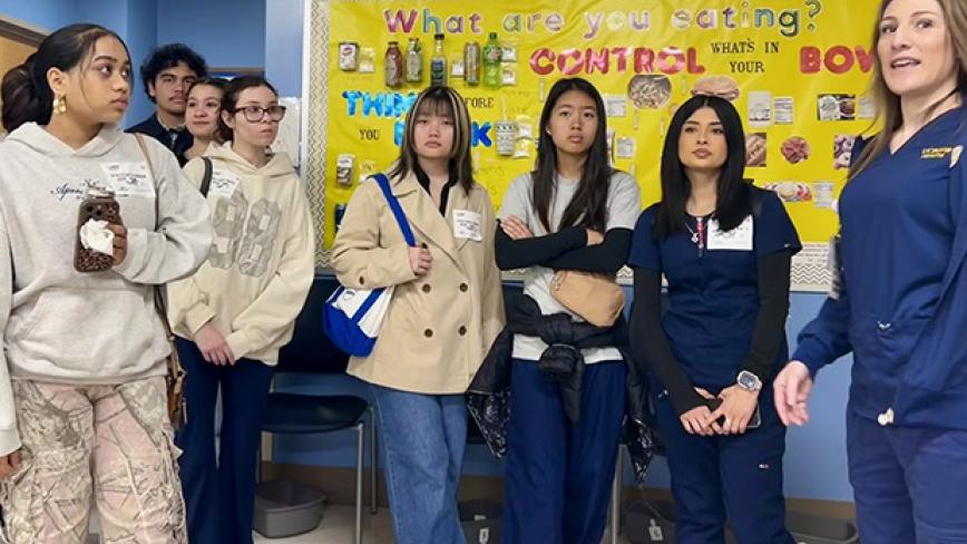 A group of high school students watch on as UC Davis Health leaders speak to them, with a board reading "what are you eating?" behind them.