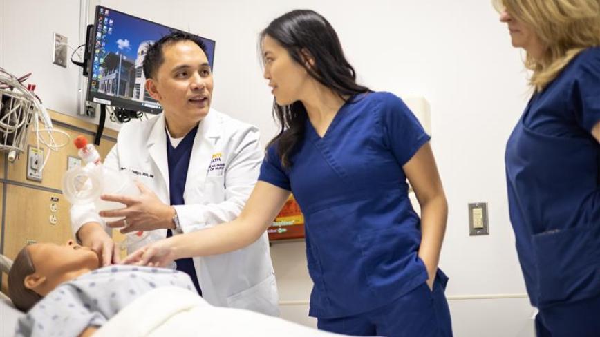 A clinical instructor in a white coat talks to two students in blue scrubs as they look at a simulation patient model in a hospital bed with a screen behind